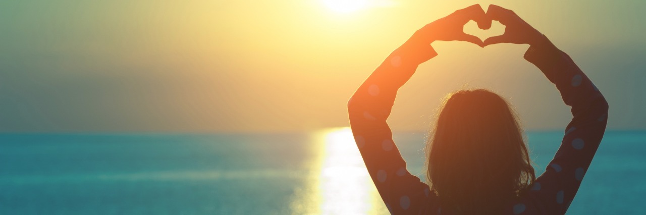 woman heart symbol hands in front of sunset over ocean