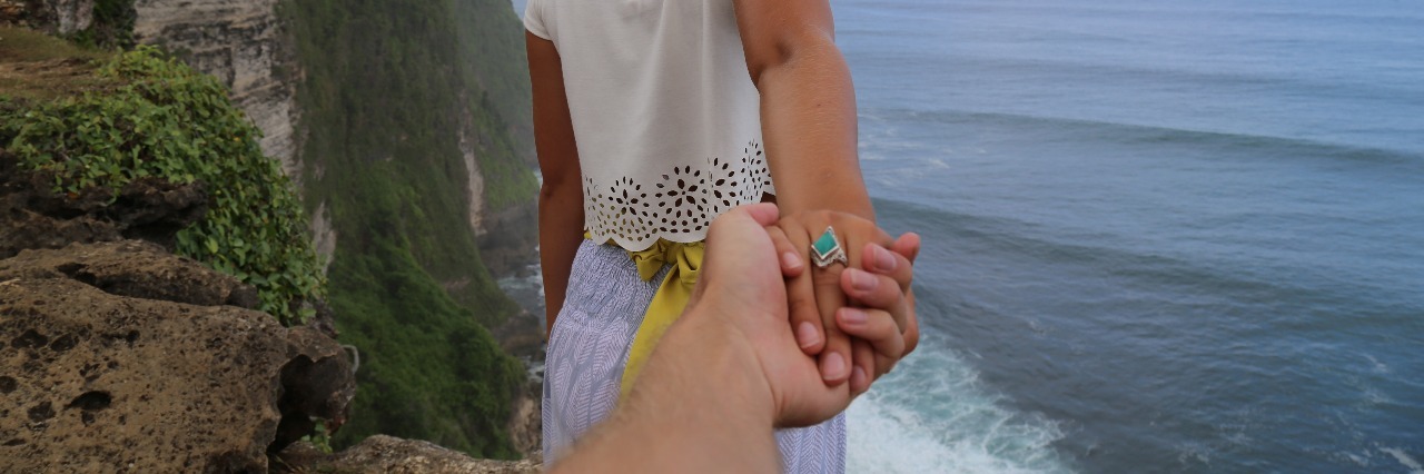 man reaching out holding woman's hand at cliff edge with view of ocean support