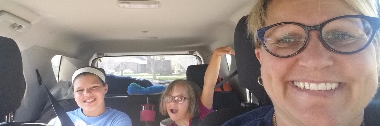 Why I Don't Identify Myself as a 'Special Needs Mom' Selfie of autor riding in the car, she has sort blond hair and wears glasses, both her daughters are sitting in the back seat and waving at the camera.