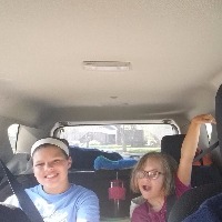Selfie of autor riding in the car, she has sort blond hair and wears glasses, both her daughters are sitting in the back seat and waving at the camera.