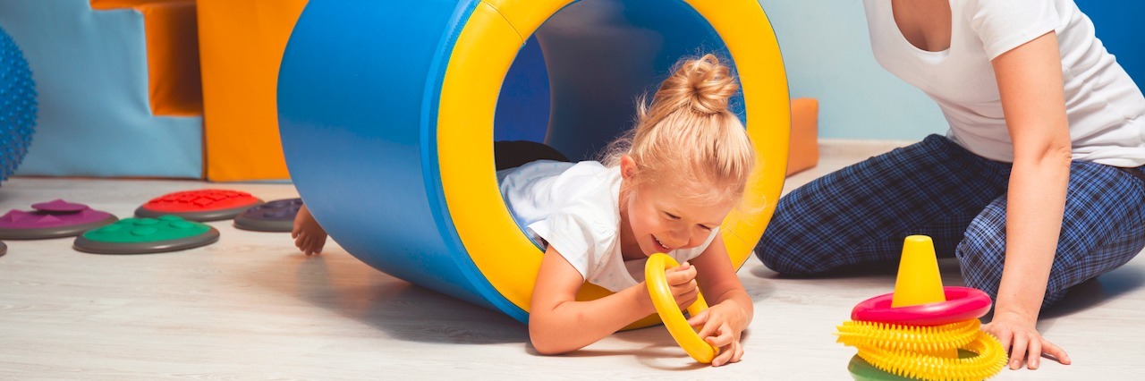 Funding for Programs That Help Kids With Physical Disabilities Matters Little girl doing therapy, she is in a roller tunnel reaching for toys while therapist keeps roller steady