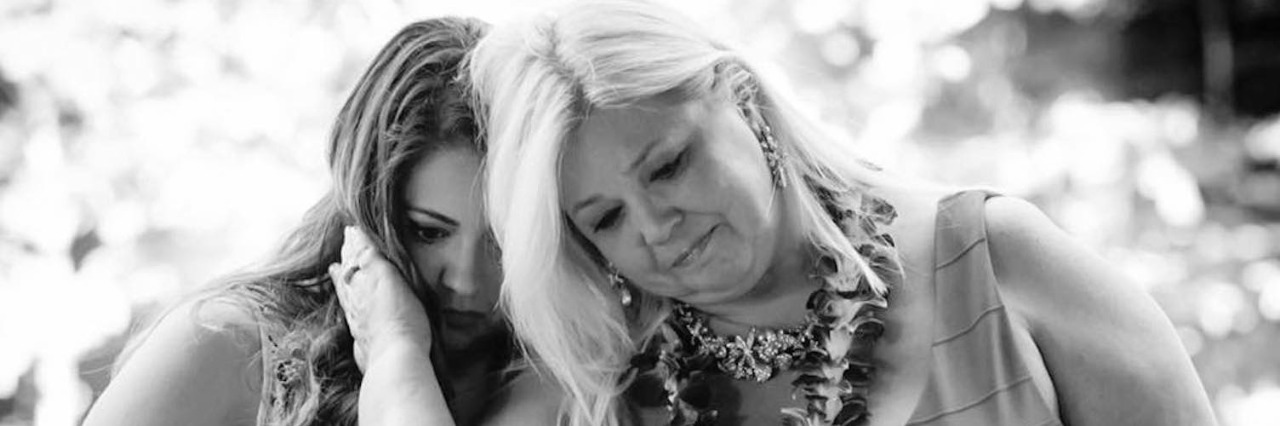Why It's Important to Talk About Grief Two women, one holding a microphone with hand on cheek of other woman, both looking down