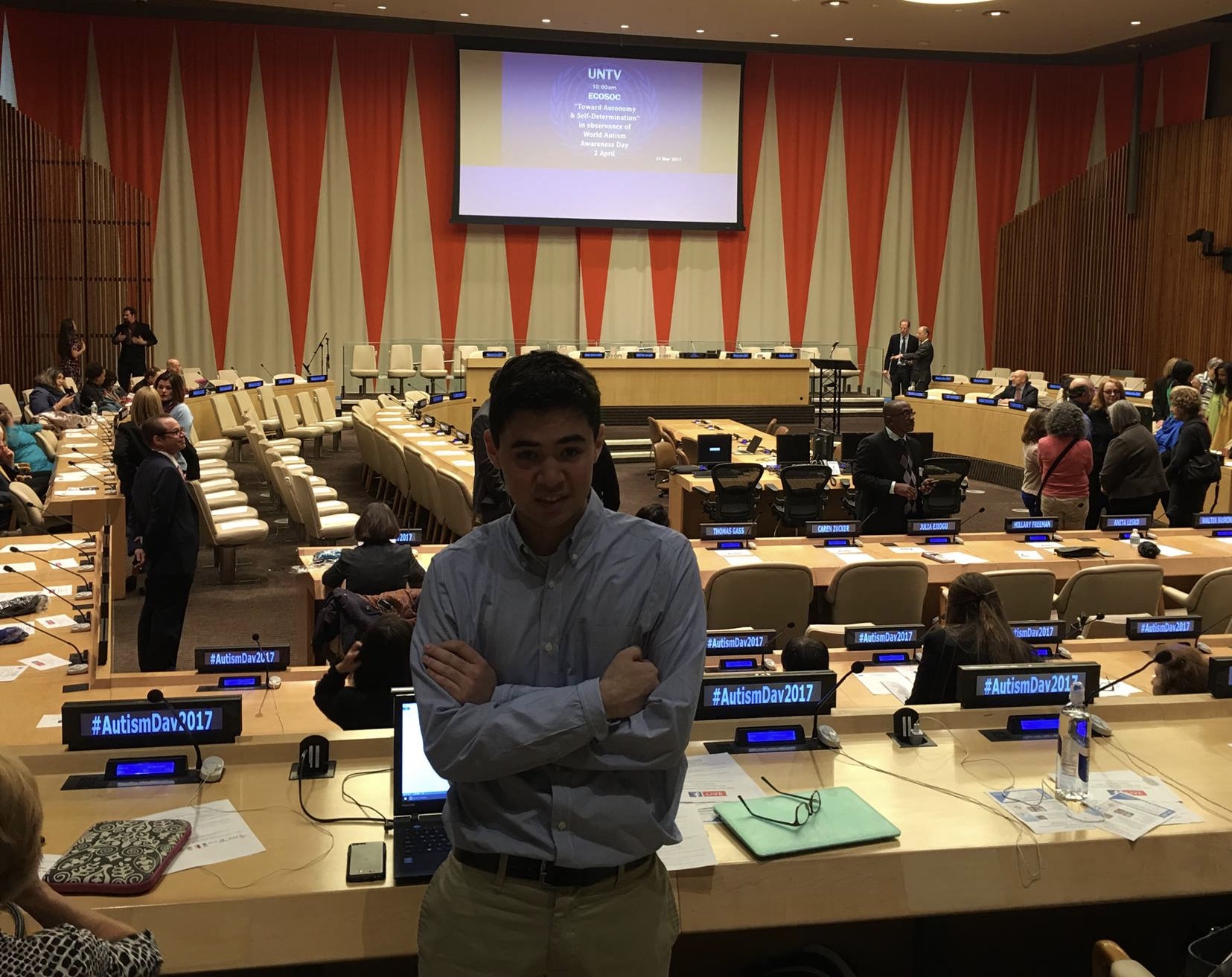 Tom at World Autism Awareness Day 2017 at the United Nations, standing in front of tables with microphones and the stage in the background