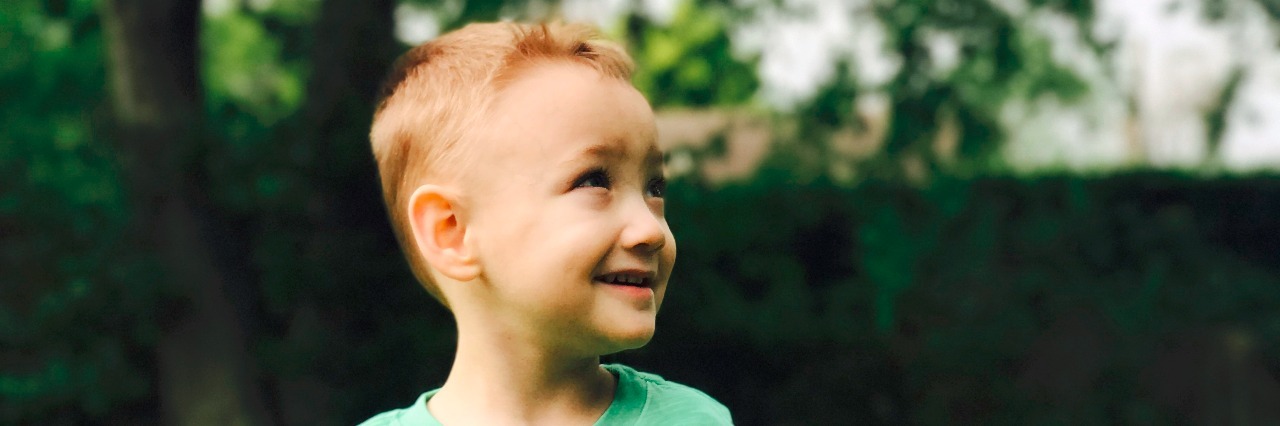 The NICU Memory That Shaped My Perception of 'Normal' Little boy standing on his backyard, wearing a green shirt, tan shorts, white socks, white leg braces, and white shoes. he is looking to the side, smiling.