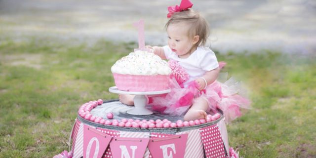 To My Daughter With Down Syndrome on Her 1st Birthday Little girl with down syndrome sitting on top of ink decorated stand and a pink birthday cake, she is reaching for the frosting