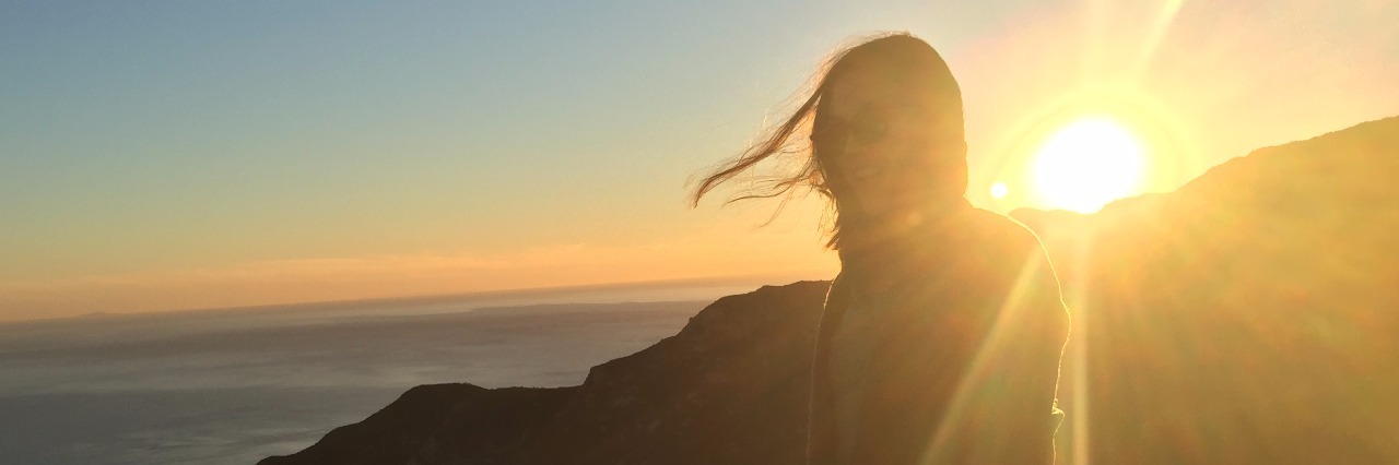 Why I Sometimes Miss My Anorexia Body image of woman standing in front of sunset smiling and looking happy with sea in background