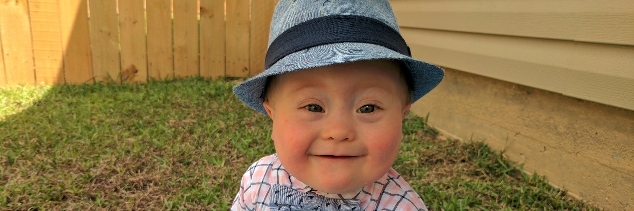 Little baby boy with Down syndrome sitting on a blue blacket outdoors. he is wearing a blue hat, plaid shirt with blue bowtie, blue jeans and blue shoes.; his Easter outfit.