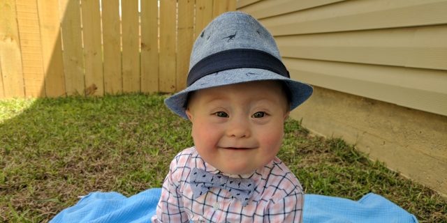 A Future Without Down Syndrome? Little baby boy with Down syndrome sitting on a blue blacket outdoors. he is wearing a blue hat, plaid shirt with blue bowtie, blue jeans and blue shoes.; his Easter outfit.