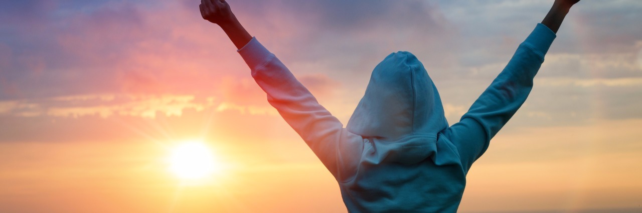 woman in blue sweatshirt raises arms victoriously in front of sunset