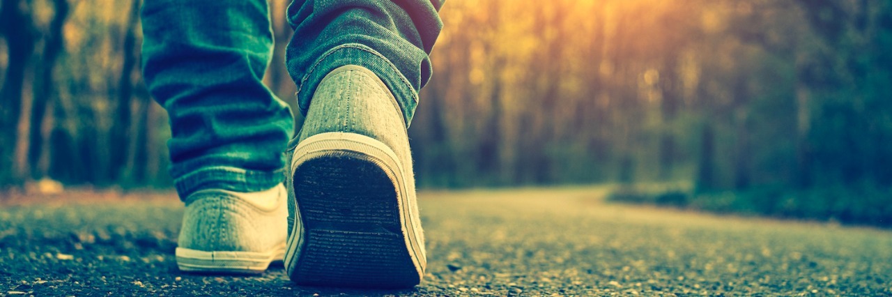 Close-up of person wearing sneakers and jeans, walking on paved path near trees