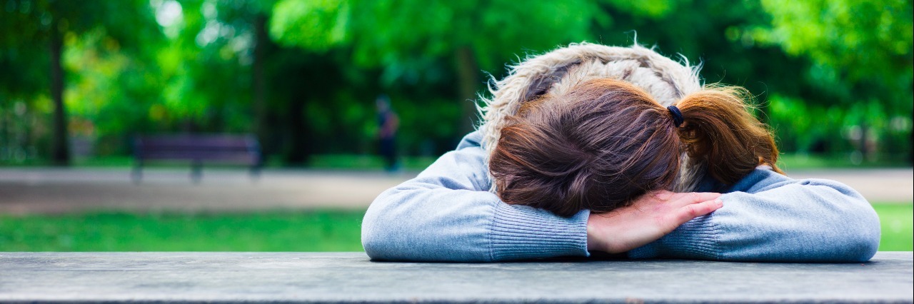 What I Really Mean When I Say 'I Need to Rest a Minute' A woman with her head resting on her hands at a table in the park