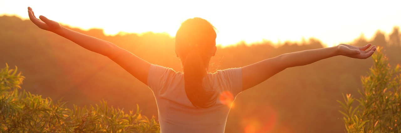 Woman standing with outstretched arms, facing sunset and tree landscape