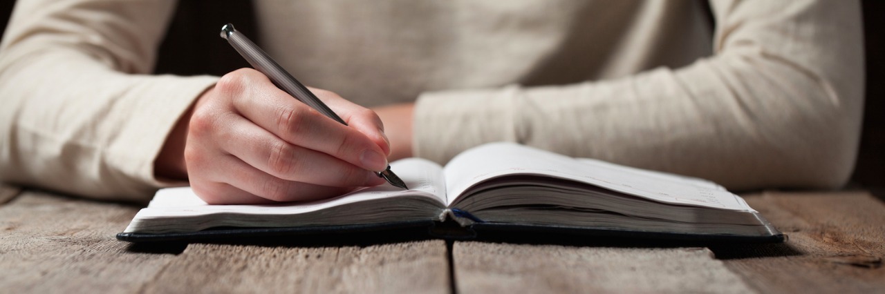 Person sitting at table with arms resting on wooden table, writing in notebook