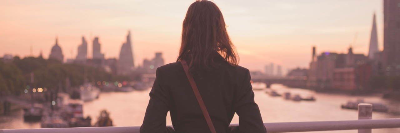 A young woman with a shoulderbag is standing on a bridge and is admiring the sunrise over the London skyline