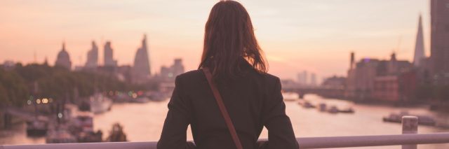 A young woman with a shoulderbag is standing on a bridge and is admiring the sunrise over the London skyline
