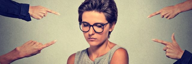 woman with glasses looking down while several hands point at her in accusation
