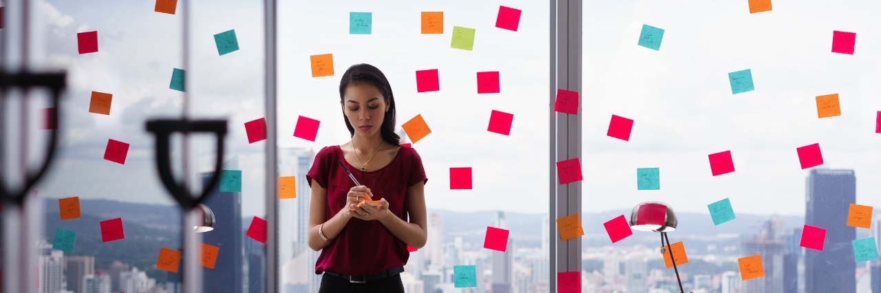 3 False Labels I've Been Assigned Because of My Chronic Illness Woman writing post it notes and sticking them on large window.