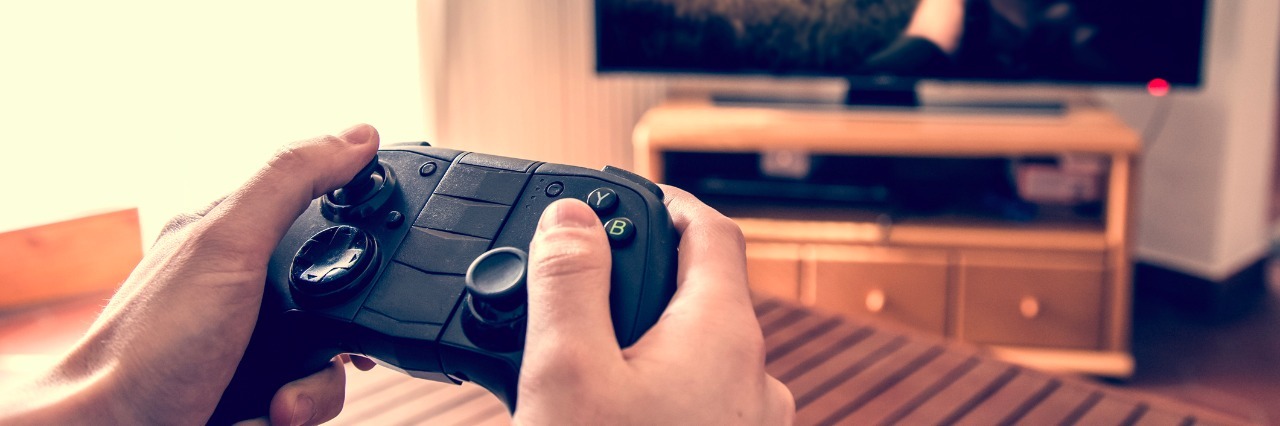 How to Use Video Games to Understand the Spoon Theory Hands holding game pad and playing shooter game on tv screen.