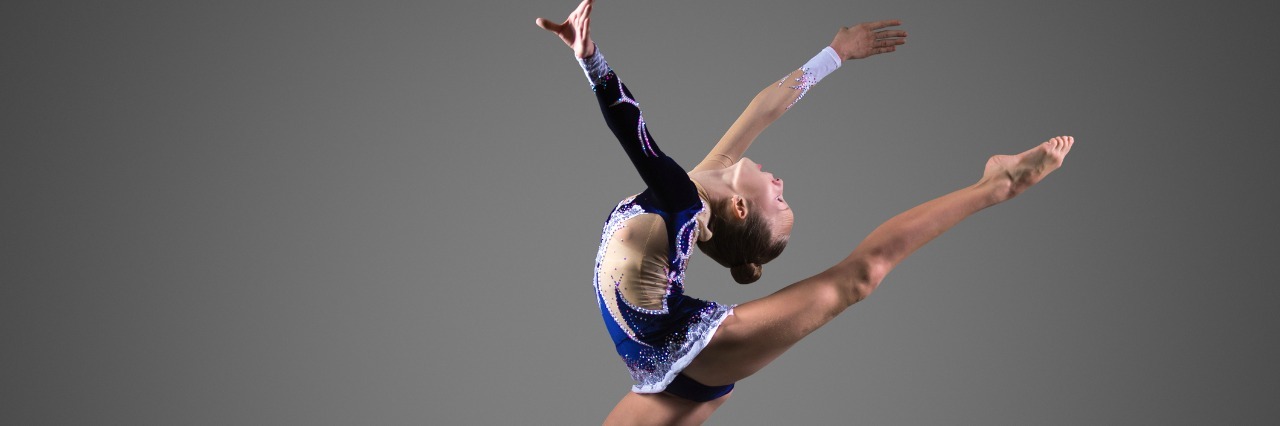 What I've Learned From Gymnastics As a Person With Mental Illness Beautiful cool young fit gymnast woman in blue sportswear dress working out, performing art gymnastics element, jumping, doing split leap in the air, dancing, full length, studio, dark background