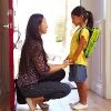 Mother and daughter in front of open door at home, with daughter wearing backpack, and mom holding daughter's hands
