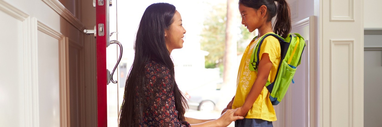 Mother and daughter in front of open door at home, with daughter wearing backpack, and mom holding daughter's hands