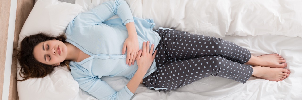 It's OK to Feel the Hard Emotions That Come With Chronic Illness Young woman laying on her back on her bed, gripping her stomach in pain.