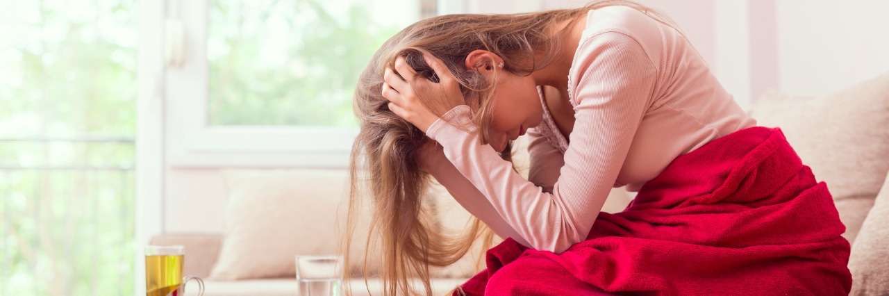 What It Really Feels Like to Live with Dysautonomia Young woman sitting on a living room couch, covered with a blanket, holding her head, having a splitting headache and pain