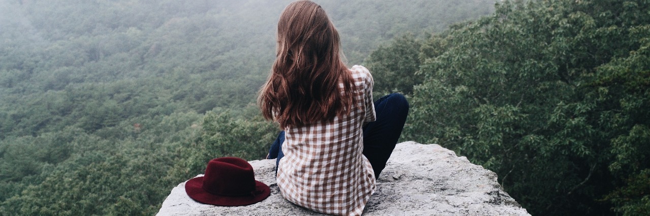 young woman sitting on cliff edge looking out over misty forest with hat by side