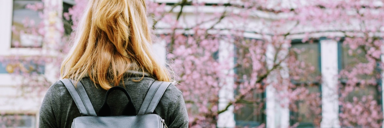 young woman in front of cherry blossoms wearing backpack facing building
