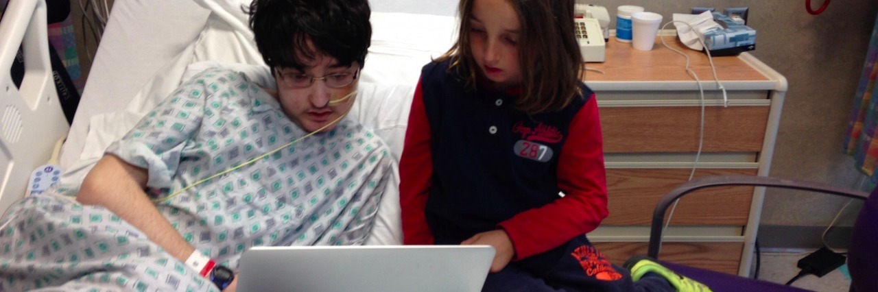 Two kids, one a hospital patient and one a visitor, sitting on hospital bed, looking at laptop