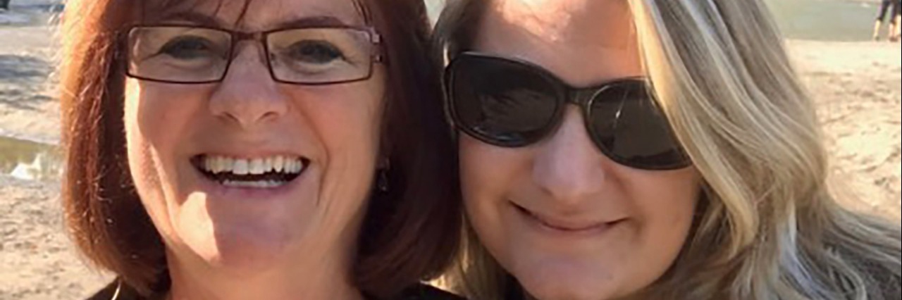 The Photograph Taken on the Day I First Considered Suicide two women in sunshine smiling with rocks and greenery behind them, one of them wearing sunglasses