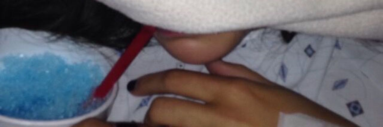 What to Know at the Beginning of Your Journey With Chronic Illness woman with her face covered by a blanket holding a blue slushie and wearing a hospital gown with iv's in her arm