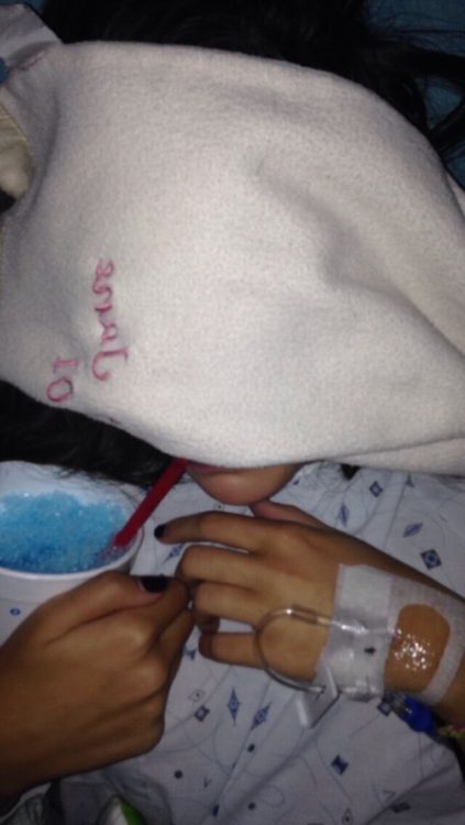 woman with her face covered by a blanket holding a blue slushie and wearing a hospital gown with iv's in her arm