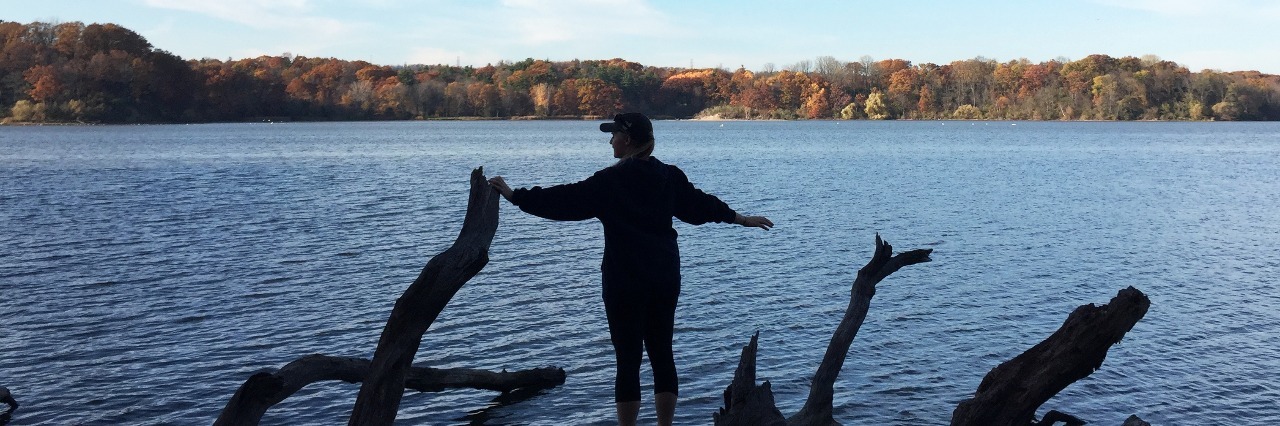 Why It's Wrong to Call Someone With Mental Illness 'Insane' young woman standing at water's edge on top of fallen tree with autumn trees in distance and blue skies
