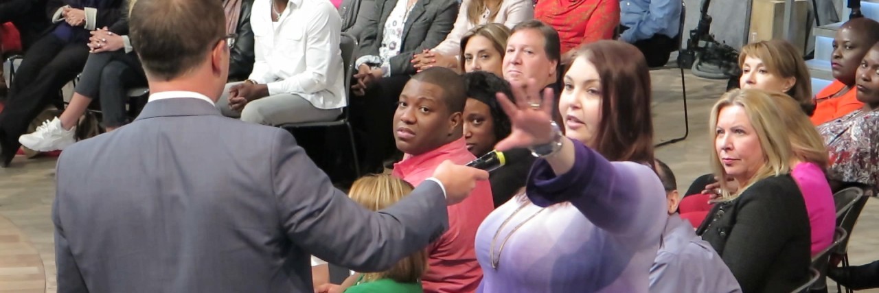 The Catch-22 Situations of Living With Fibromyalgia and Sarcoidosis A talk show host holds a microphone toward a woman in a purple top seated in the audience, as she raises her hand to speak while other audience members look on.