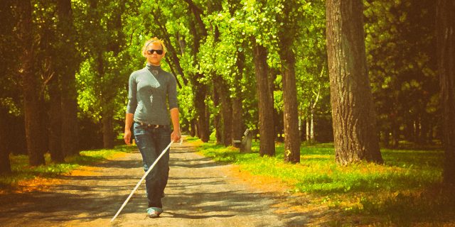 A woman using a white cane walks in a park.