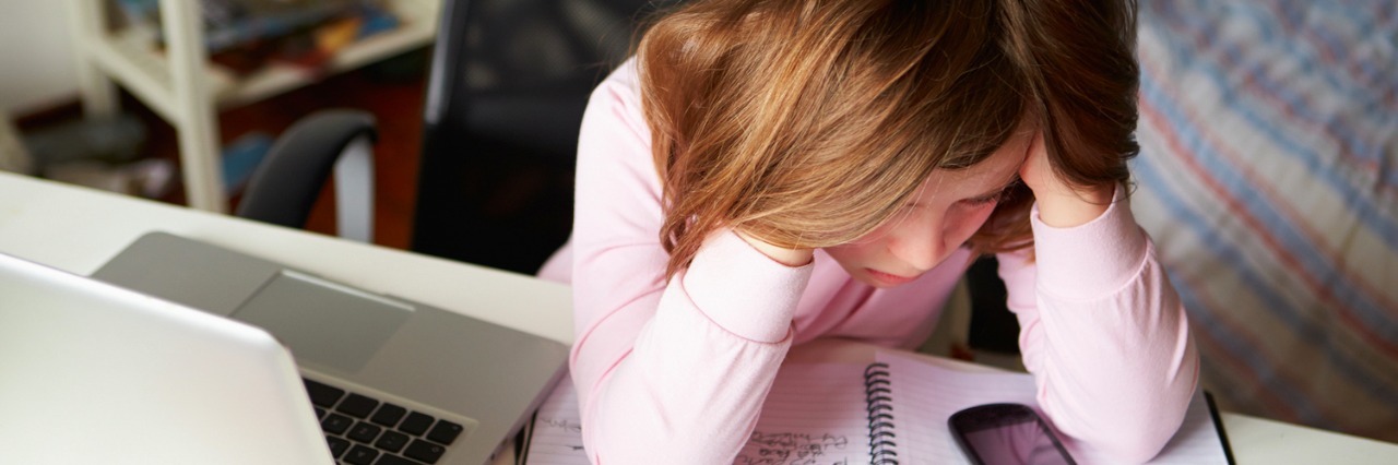 Girl with hands on head, sitting at desk with laptop, cell phone and notebook