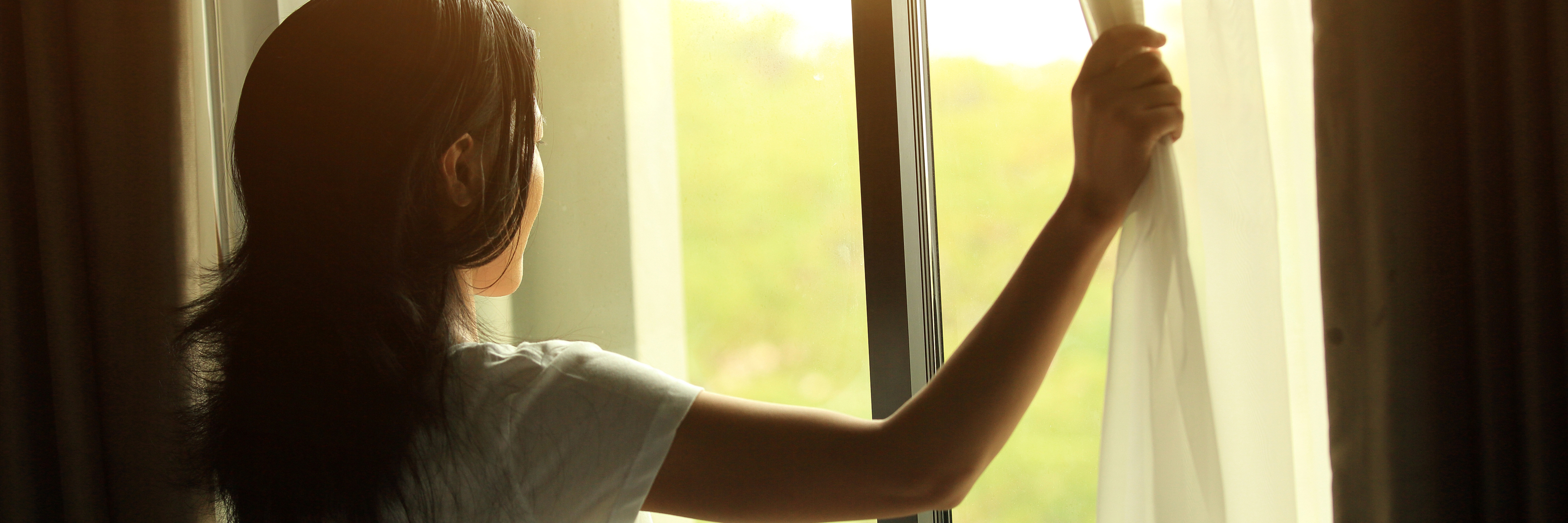 The Truth About Self-Care When It Seems Like a Struggle young woman opening curtains in bedroom letting light in