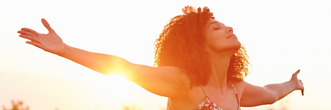 woman standing outside with her arms outstretched and the sun setting behind her