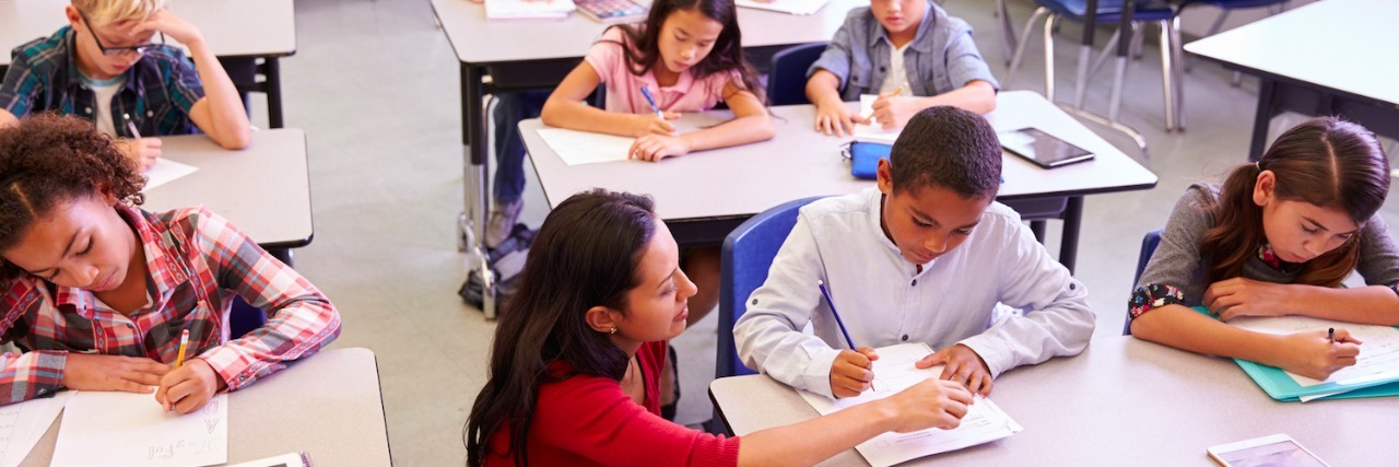 Teacher next to student's desk in class of elementary school kids writing at their desks