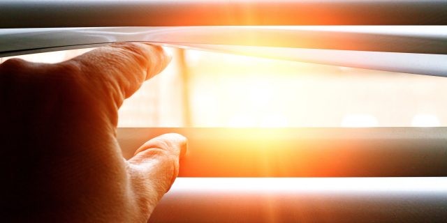 Man looks through blinds, seeing sunlight.