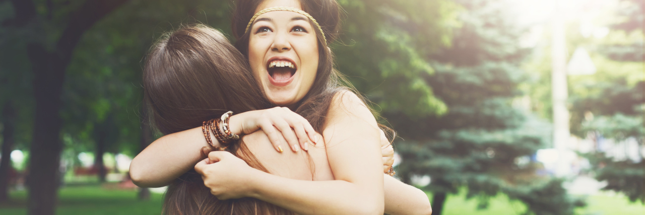 Two happy young girls hug each other. Females embracing, laughing and excited. Woman friendship, walk in the park outdoors. Asian boho girl with friend