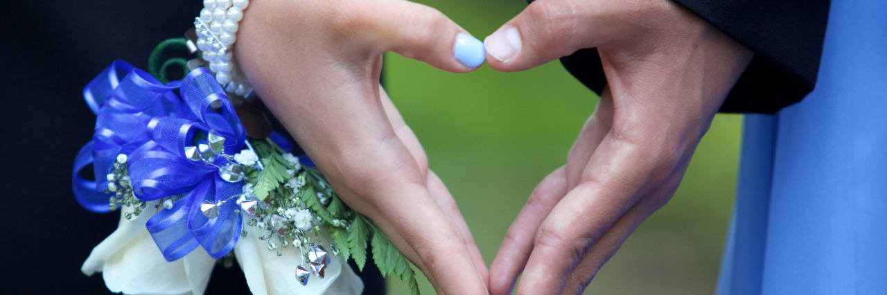 A couple going to the prom forming a heart shape with their hands. She is wearing a blue and white wrist corsage.