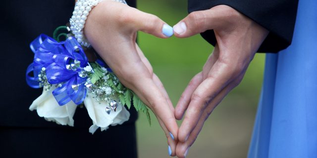A couple going to the prom forming a heart shape with their hands. She is wearing a blue and white wrist corsage.