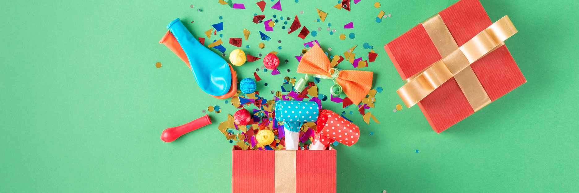 Red gift box with various party confetti, balloons, streamers, noisemakers and decoration on a green background. Colorful celebration background. Flat lay.