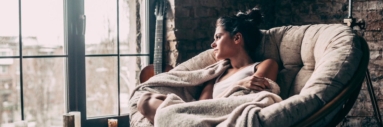 What It Means to Say 'If You Need Anything, I'm Here' to a Sick Friend woman sitting on chair by window covered in blanket