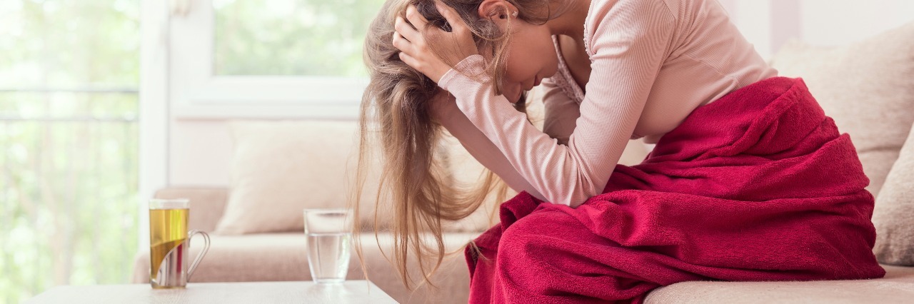 young woman sitting on couch with a blanket holding her head in pain