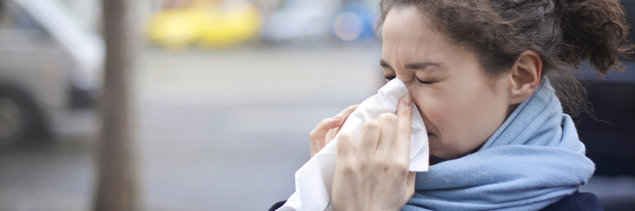 Asthma, Arthritis, Fibromyalgia: Getting a Cold With Chronic Illness “Person wrapped in a scarf blowing their nose outdoors, showing symptoms of a cold.”