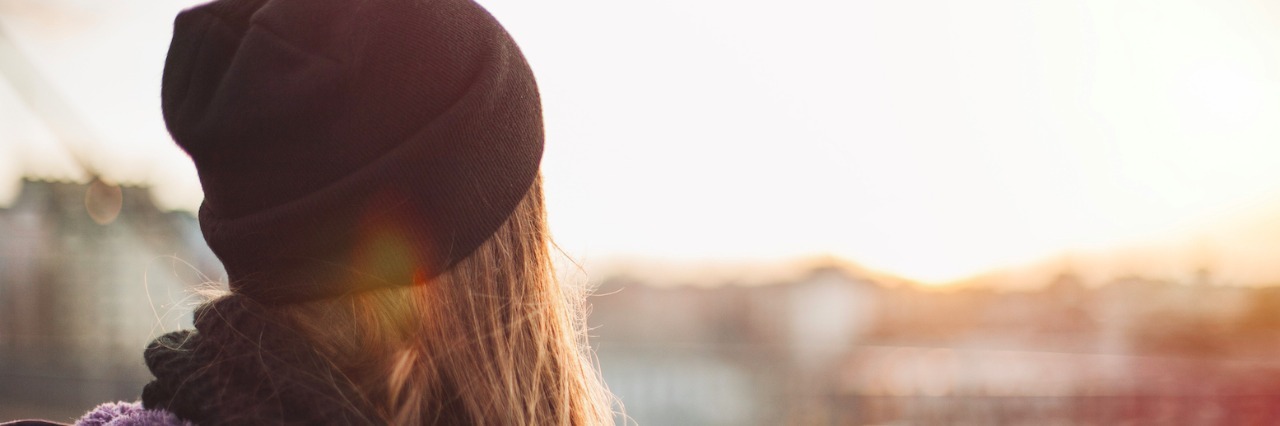 My Autism and Being Unable to Speak at Times Woman wearing beanie and hoodie, on city street and facing building landscape and sunset sky