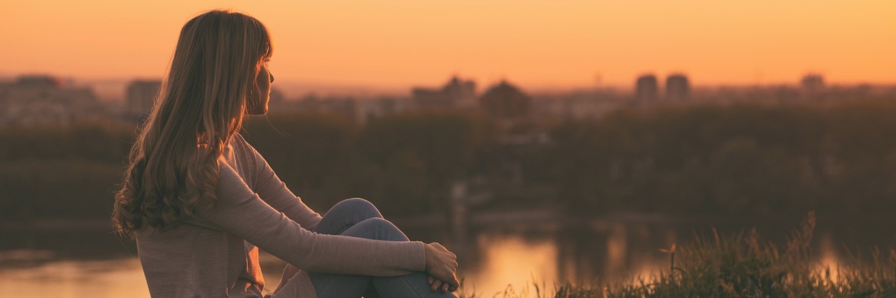 When People Think Your Life Looks Easy With Chronic Illness woman sitting on a hill overlooking a city during sunset